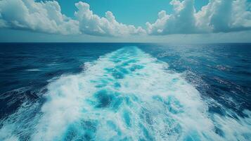 Ocean wake pattern stretches behind cruise ship in aerial view. Deep blue sea meets bright sky with white clouds, while turquoise foam trails create dynamic water movement on sunny day. maritime photo