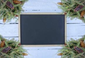 Christmas composition with fir branches and cones on white aged wooden background. New Year festive design template. Empty framed chalkboard. photo