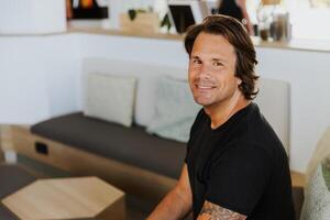Portrait of a smiling middle aged man with tattoos on his arm and long hair sitting on the couch photo