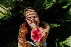 Smiling woman holding a pink flower while sitting on a grass field. Shadow of leafs on her face photo