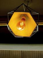 Light bulb is lit up in a hexagonal shaped lamp. The light is yellow and the lamp is hanging from the ceiling photo