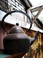 Black tea kettle sits on a wooden shelf. The kettle is old and rusted, and it has a chain hanging from it. The scene is somewhat eerie and gives off a sense of nostalgia photo