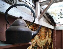 Black tea kettle sits on a wooden shelf. The kettle is old and rusted, and it has a chain hanging from it. photo
