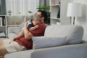 Side view shot, Single father in a casual striped polo shirt sits on a sofa holding his young son and lulling him to sleep in a cozy room at home. photo