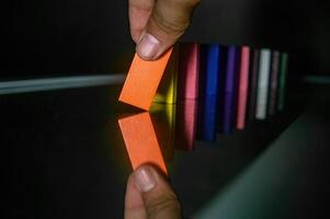 A hand is seen tipping an orange domino, initiating a chain reaction with other colorful dominoes lined up on a reflective surface. The image captures the moment of action and reflection photo