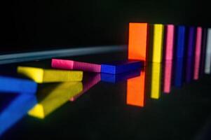 Colorful dominoes in a line, with some fallen over, reflecting on a shiny surface. The vibrant colors and reflections create a visually striking and dynamic composition photo