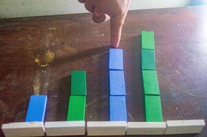 A hand points to a colorful bar chart made of wooden blocks on a wooden table, illustrating data comparison with green and blue blocks of varying heights photo