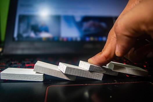 A close up of a hand pushing over a row of white dominoes on a laptop keyboard, illustrating the concept of cause and effect or chain reaction in a digital workspace photo