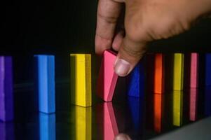A hand is seen tipping over a row of colorful dominoes, including yellow, pink, blue, and orange pieces, creating a chain reaction effect photo