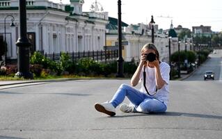 The image shows a woman holding a professional DSLR camera, positioned against a backdrop of a building with a stone facade. photo