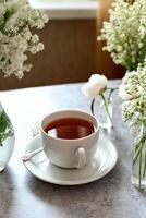 A white teacup rests on a matching saucer with a small spoon, surrounded by vases of delicate white and green flowers, all set against a mottled grey wall. photo