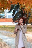A woman with auburn hair, dressed in a tan trench coat, is talking on a smartphone, holding brown-tinted sunglasses in her left hand. photo