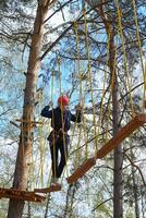 A medium shot featuring a person walking across a wooden beam, with a complex web of ropes and trees surrounding them, under a bright blue sky photo