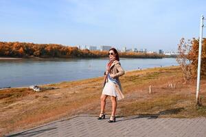 A full-length shot of a woman with a scarf, set against a background of a river, trees, and a blue sky, using a diagonal line of sight. photo