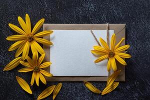 A flat lay image featuring yellow flowers arranged around a blank card and envelope against a dark, textured surface photo
