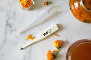 A flat lay image of a digital thermometer, calendula flowers and petals, and a glass jar on a white marble surface, creating a wellness or herbal remedy scene. photo