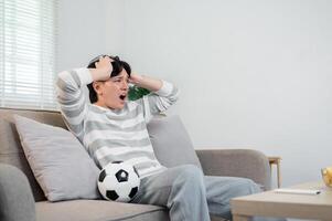 A shocked Asian man is watching his favorite football team lose to another team on TV at home. photo