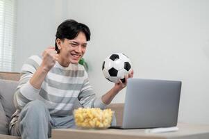 A happy Asian man enjoys watching his favorite football team's match via his laptop at home. photo