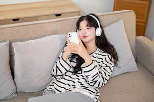 A charming Asian woman is relaxing on a sofa, chatting with her friends on her smartphone. photo