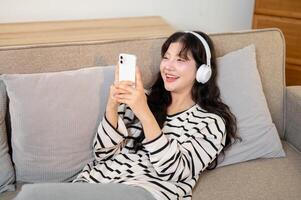 A charming Asian woman is relaxing on a sofa, enjoying chatting with her friends on her smartphone. photo