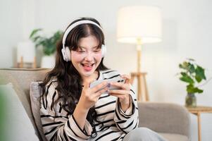 A happy Asian woman wearing headphones enjoys watching a game show on her smartphone on a sofa. photo