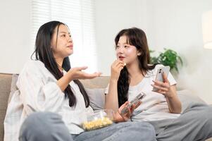 Two happy Asian female friends sit on a sofa, enjoying snacks and sharing their stories. photo