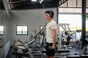 Focused Asian man is working out with dumbbells in front of the dumbbell rack and mirror in the gym. photo