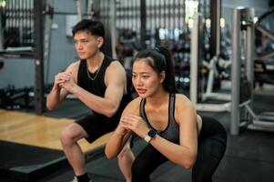 Side view of a sporty Asian couple in sportswear is training together in the gym, performing squats photo