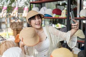 A cheerful Asian-Chinese female customer enjoys blogging on her phone while shopping in a hat shop. photo