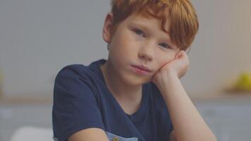 A boy with ginger hair sits at a table, resting his chin on his hand while concentrating on his schoolwork. He is participating in a remote lesson, focused and dedicated to learning. video