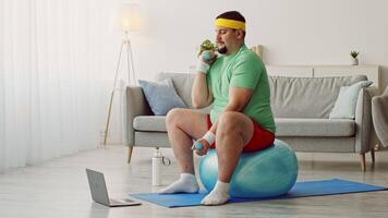 A chubby millennial man is working out at home by sitting on an inflatable exercise ball. He is holding dumbbells and following a workout routine on his tablet in a bright living room. video