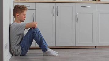 A young boy sits on the floor against the wall in a kitchen, showing signs of sadness and isolation. The environment appears tidy, emphasizing his emotional state. video