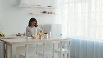A millennial African American woman joyfully prepares baked goods in her cozy kitchen. The well-lit space features simple decor, highlighting her cooking activities and love for food. video