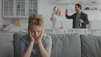 A child sits on the couch with hands over ears, visibly distressed by the ongoing argument between parents in the kitchen. Emotions run high in this unsettling domestic environment. video