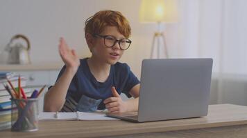 A boy with ginger hair is engaged in his online studies. He sits at a desk with a laptop, concentrating on his lesson. The room is bright and organized, supporting his learning environment. video