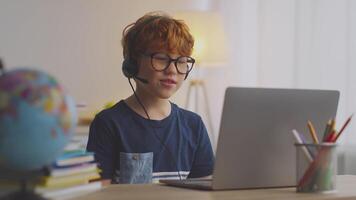 A preteen boy with ginger hair is focused on his laptop during a remote learning session. Surrounded by school supplies, he wears headphones, indicating active participation in lessons. video