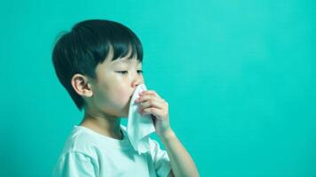 A boy holds a tissue to his nose, sneezing against a turquoise background photo