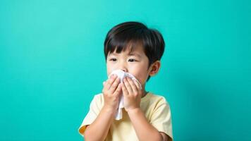 A boy sneezes into a tissue with a tissue box nearby against a turquoise back photo