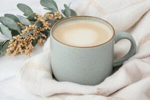 Cozy beige mug with frothy latte on soft cloth with eucalyptus and yellow flowers in the background. Cozy vibes concept. photo