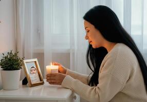 Woman lighting memorial candle beside baby's photo on table in cozy room