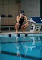 Female swimmer preparing to dive at indoor poolside in black swimsuit and goggles photo