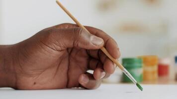 A young African American man focuses on painting, holding a brush dipped in green paint. The setting features various paint containers and a blank canvas, showcasing a moment of artistic expression. video