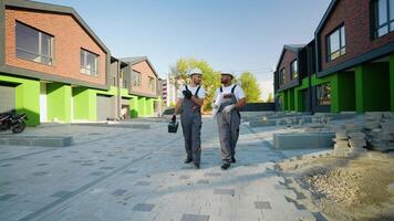 Two multiethnic builders in hard hats and workwear walk near a modern green and brick townhouse, discussing the progress of the project video