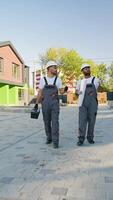 Two builders in hard hats and workwear walk near a modern green and brick townhouse, discussing the progress of the project video