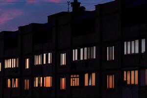 A multi-story brick building at dusk with illuminated and dark windows. Antennas and rooftop structures are visible. The sky transitions from deep blue to soft pink, creating a twilight ambiance. photo
