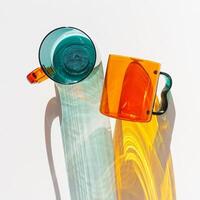 Vibrant still life color transparent glass mugs or cup in shades of green teal and amber orange. Sunlight dynamic shadow and reflections on white, minimal aesthetic design, top view summer drink photo