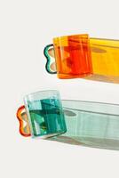 Two Modern Glass Cups with Vibrant sunlight Shadows, Aesthetic Top view of transparent orange and teal color glass mugs for drink. Yellow green reflections, shadow on white, minimal lifestyle design photo