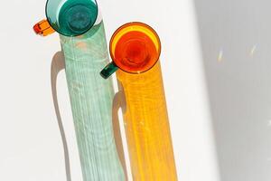 Vibrant still life colorful transparent glass mugs or cup in shades of green teal and amber orange. Sunlight dynamic shadow and reflections on white, minimal aesthetic design, top view summer drink photo