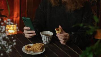 Tea time, daily routine. Woman using mobile phone sitting at table with cup of tea and pastries in cozy room. video