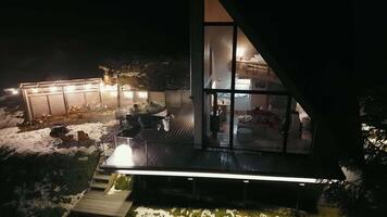 Nighttime view of modern mountain cabin with hot tub, A modern A-frame mountain cabin with warm lights is surrounded by snow video